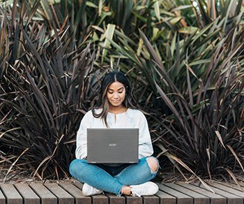 A person sits on a wooden walkway with lots of vegetation in the background. They are sitting forward-facing with legs crossed and a laptop balanced on their legs.