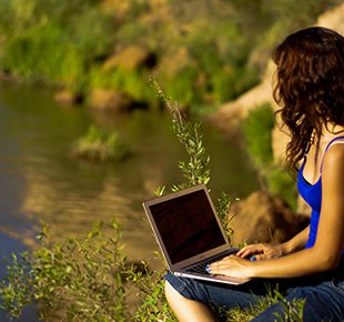A person with long hair sits outside with an open lap top next to a river.