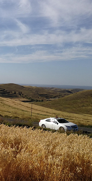 A white car drives on a road with mountain and agricultural scenery in the background